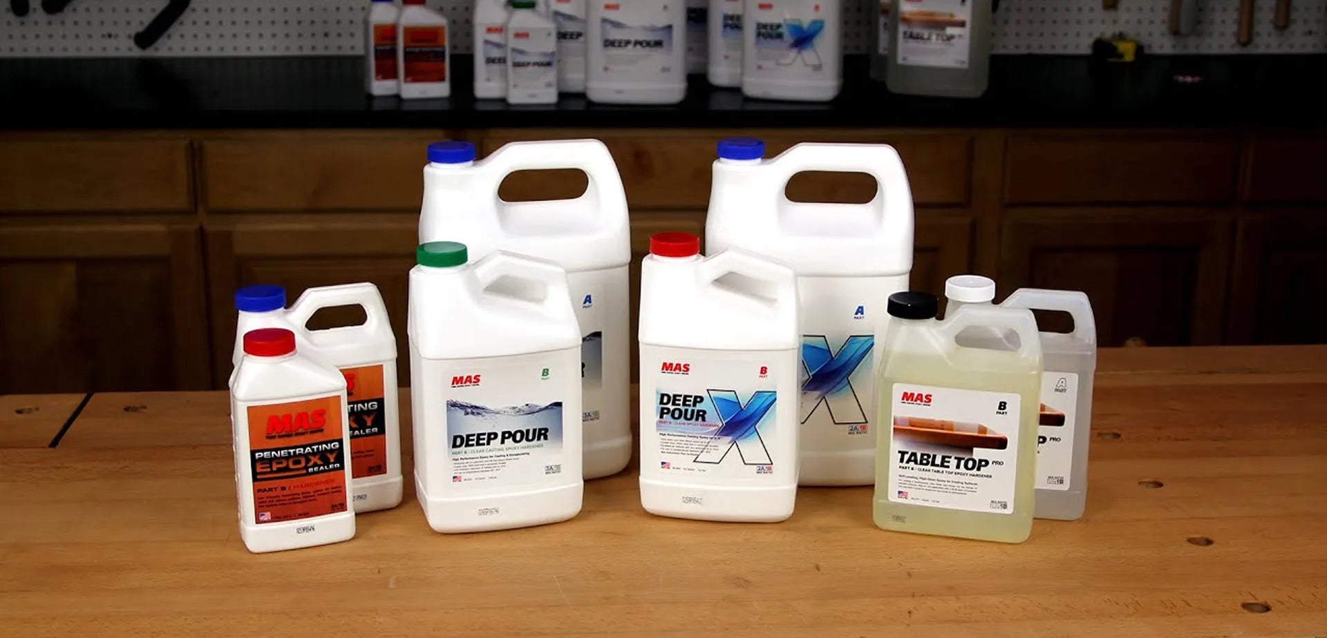 Lineup of MAS Epoxies product bottles, including Penetrating Epoxy Sealer, Deep Pour, Deep Pour X, and Table Top Pro epoxy, arranged on a wooden workbench in a workshop